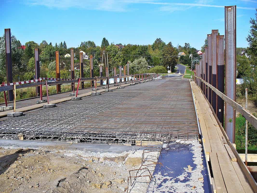 The framework of a new bridge under construction, showcasing extensive rebar reinforcement set against the backdrop of a clear blue sky. Steel beams rise vertically along the sides, and the road extends into a rural landscape, indicating a blend of infrastructure development within a natural setting.