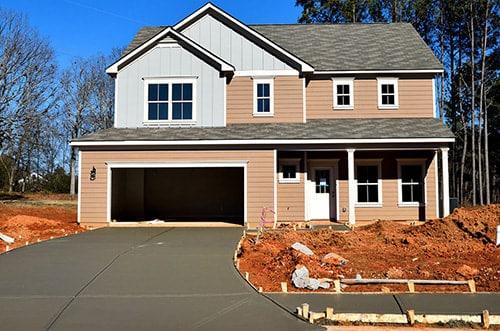 A newly constructed two-story house with beige siding and a gray roof, featuring an open garage and a freshly poured concrete driveway. The surrounding area is unfinished with exposed red clay soil and wooden formwork, indicating ongoing landscaping or construction work under a clear blue sky.
