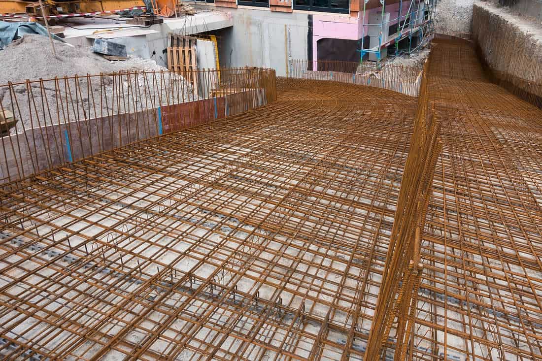 An expansive grid of rusty rebar laid out for a large concrete foundation, with a construction site in the background featuring piles of gravel, scaffolding, and partially constructed walls. This structural steel reinforcement is prepared for a concrete pour, indicative of an early stage in a building's construction.