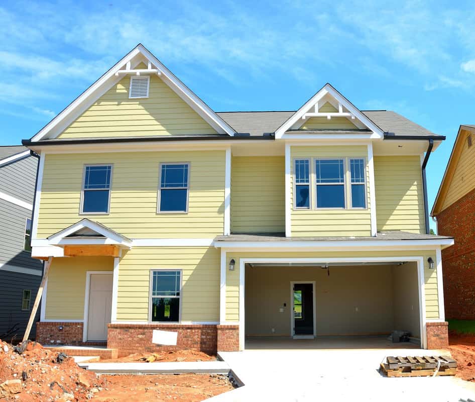 A new two-story home with pale yellow siding and white trim, featuring an open two-car garage and unfinished front steps. The house, under clear blue skies, awaits further exterior finishes, as evidenced by the surrounding construction materials and bare red clay soil.