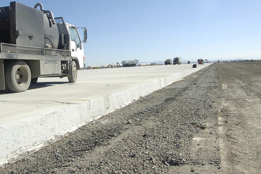 A road construction scene with a white and black street sweeper truck on the left and freshly laid concrete curbs on the right. The background shows a clear sky and distant construction vehicles and equipment along the length of the road under construction.