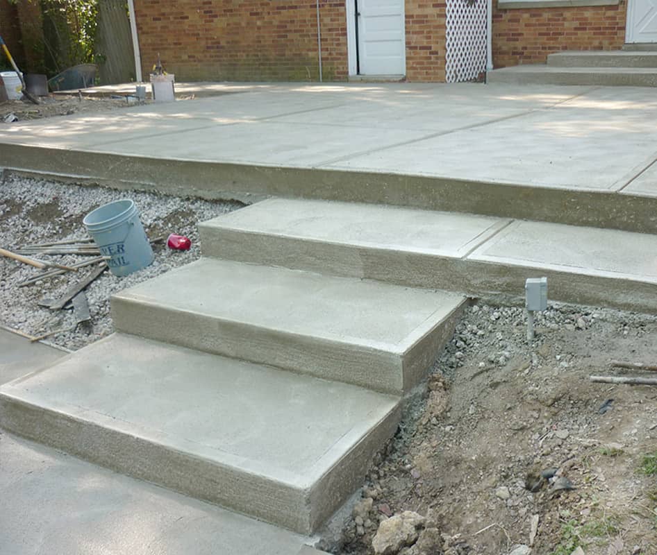 Newly constructed concrete steps lead to a patio area adjacent to a brick house with a white garage door. The scene is a work in progress, with construction debris, a blue bucket, and wooden planks scattered around the site, awaiting cleanup and final touches.