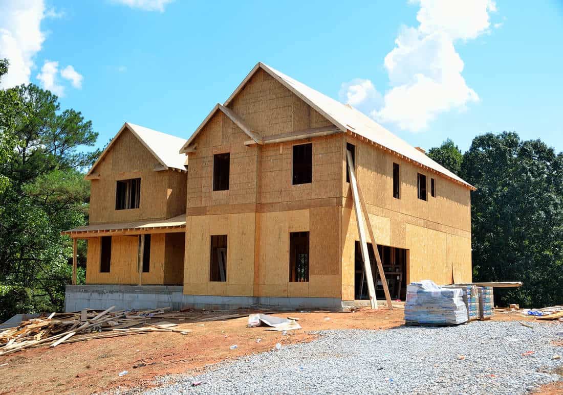 A new residential construction with wooden framing and a covered porch in progress. The structure, covered in tan sheathing, stands under a clear blue sky with a backdrop of dense green trees, with construction materials and debris scattered in the foreground.