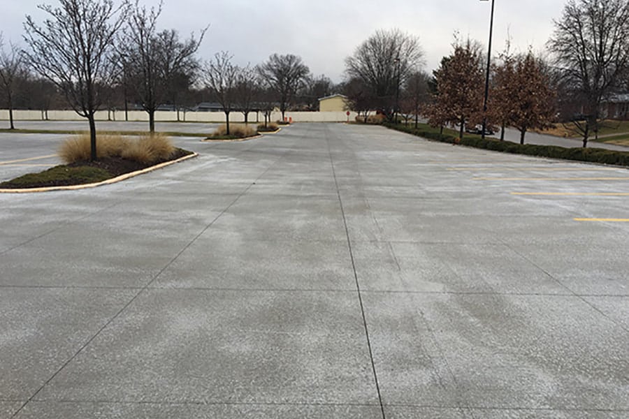 An expansive parking lot with freshly painted yellow parking space lines. The overcast sky suggests a chilly or damp day. Surrounding the parking lot are winter-bare trees and well-maintained landscaping with ornamental grasses.