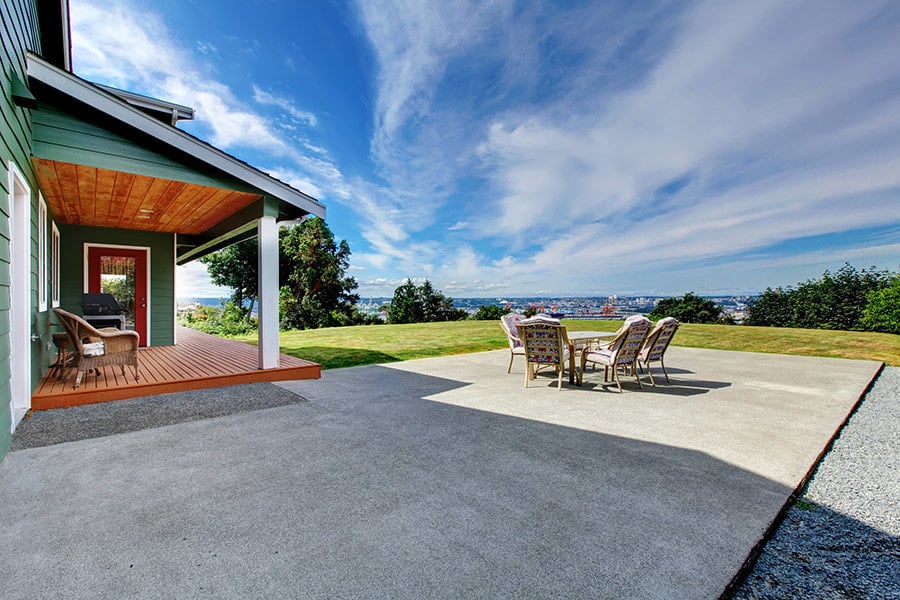 A spacious concrete patio extends from a house with a covered wooden deck, leading to an outdoor dining area with a table and chairs set against a breathtaking view of a distant city skyline beneath a vast, cloud-streaked sky.