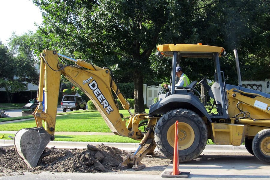 A construction worker operates a yellow John Deere 310C backhoe loader near a roadside, with the backhoe arm positioned over a small pile of dirt. An orange traffic cone is placed in front of the machine for safety. Residential homes and lush greenery provide a suburban backdrop to the work site.