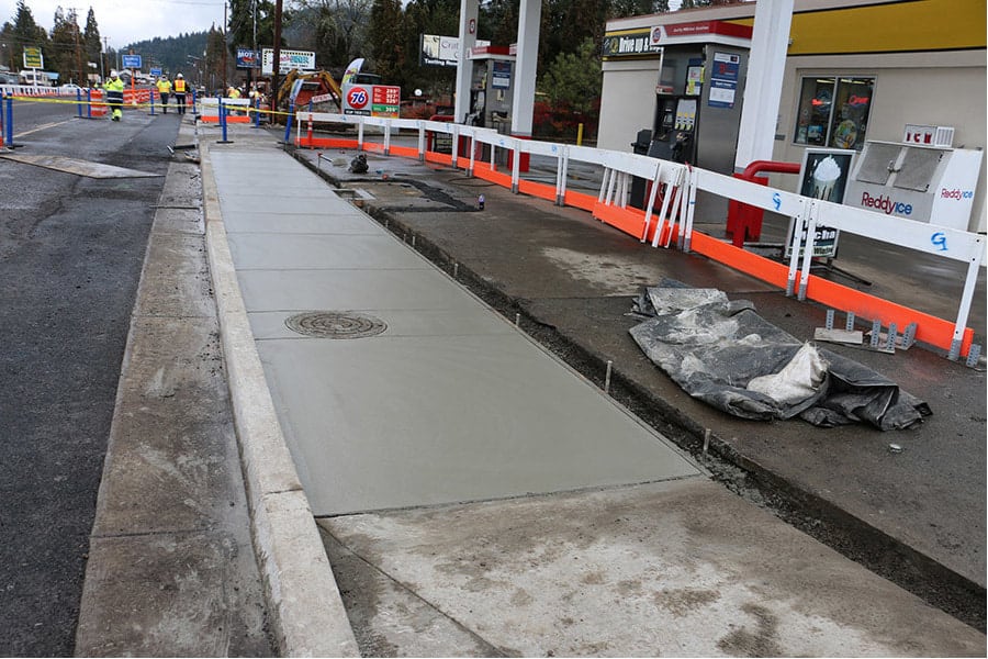 A newly poured concrete sidewalk with a manhole cover, framed by an orange safety barrier at a construction site. A gas station with a prominent 76 logo is visible in the background, with overcast skies suggesting recent or impending work on the pavement.