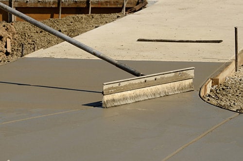 A bull float is used on a fresh concrete surface to ensure it is smooth and level. The long handle of the tool extends diagonally across the photo, showing the process of finishing the concrete. In the background, construction forms and a gravel substrate are visible, indicating an active construction site.