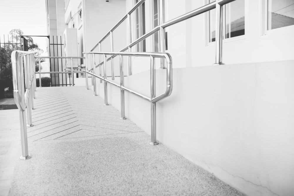 A grayscale image of a ramp with textured paving and stainless steel handrails leading to a building entrance. The picture, taken from a low angle, shows the accessibility feature in a modern architectural context, with a focus on the handrails and the edge of the ramp.