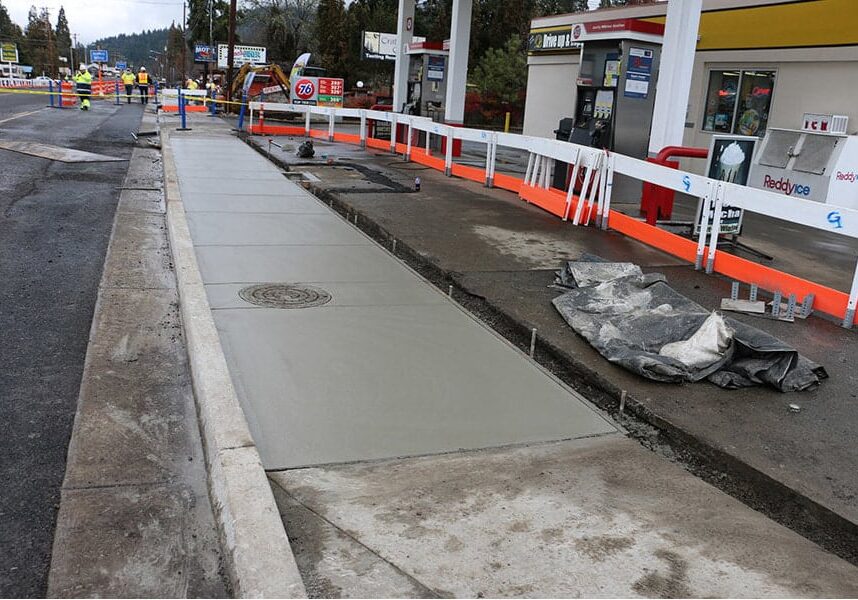 A newly poured concrete sidewalk with a manhole cover, framed by an orange safety barrier at a construction site. A gas station with a prominent 76 logo is visible in the background, with overcast skies suggesting recent or impending work on the pavement.